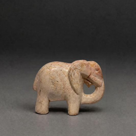 A small carved elephant figurine in a light beige stone or resin with subtle brown marbling, photographed against a dark gray studio background.