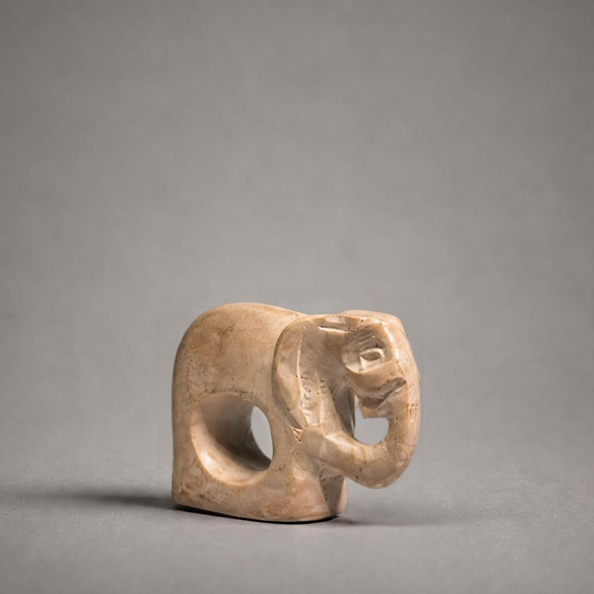 A small carved elephant figurine in a light beige stone or ceramic material, positioned against a neutral gray studio background.