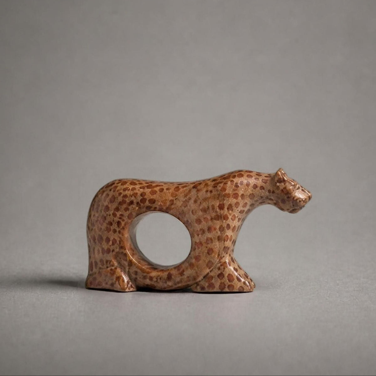 A small carved figurine of a cheetah with a circular cutout in its body, made from brown spotted wood, set against a solid gray background.