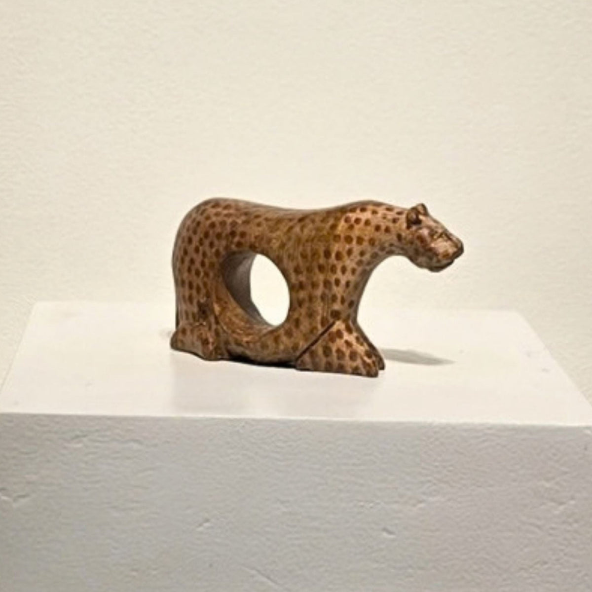 A small carved wooden cheetah figurine with a circular cutout in its side, displayed on a white pedestal against a plain off-white wall.