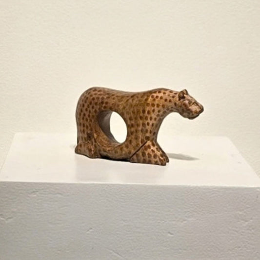 A small carved wooden cheetah figurine with a circular cutout in its side, displayed on a white pedestal against a plain off-white wall.