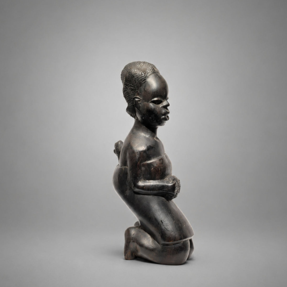 A dark wood sculpture of a kneeling child in profile, shown against a neutral gray studio background.