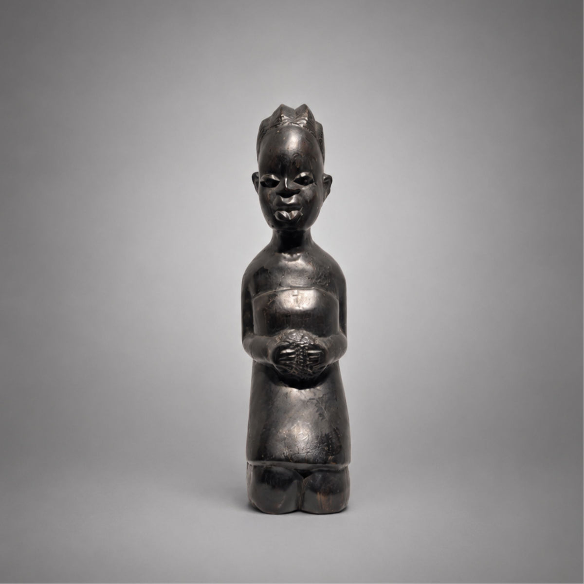 A dark ebony wooden figurine of a kneeling child, photographed against a smooth gray studio background.