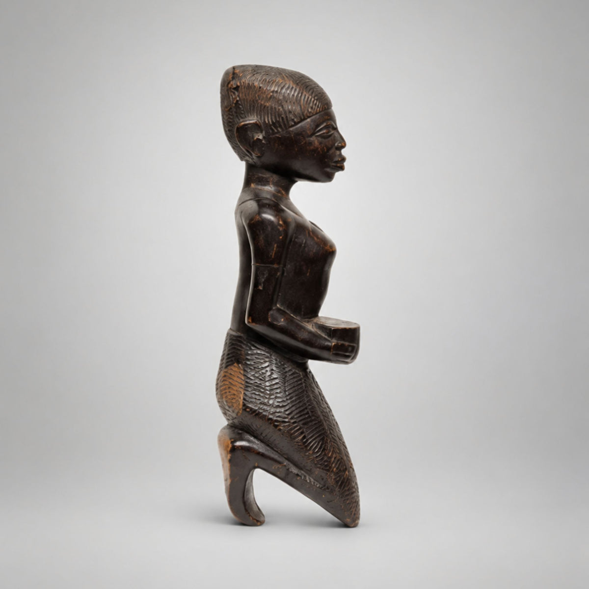 A dark carved wooden figurine of a kneeling human figure in profile, photographed against a plain light gray studio background.