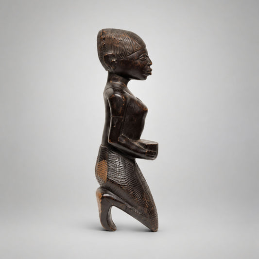 A dark carved wooden figurine of a kneeling human figure in profile, photographed against a plain light gray studio background.