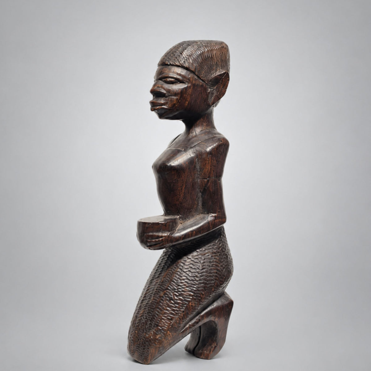 A dark brown carved wooden figurine of a kneeling female figure with textured details, shown against a neutral light gray background.