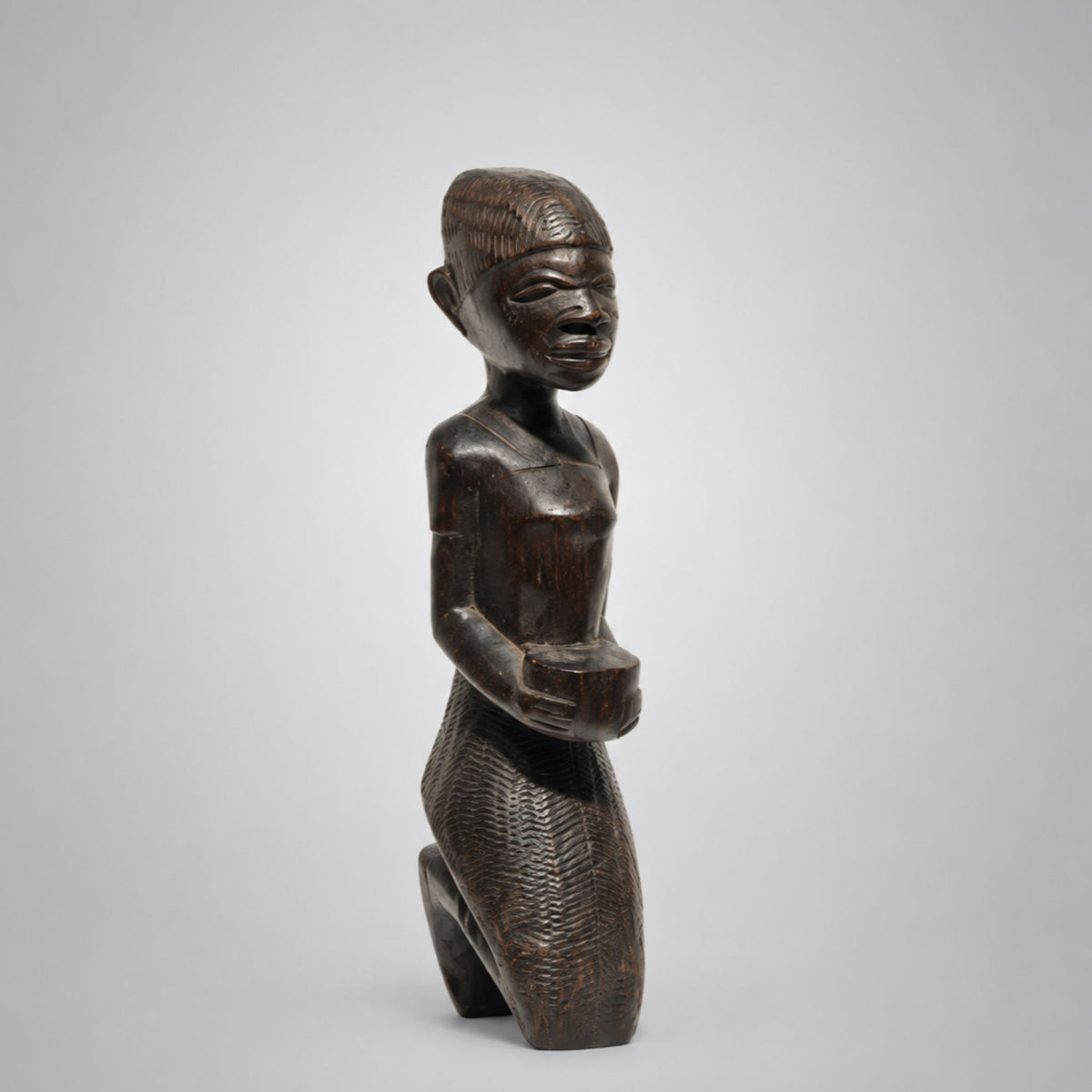 A dark brown carved wooden figurine of a kneeling figure with a stylized face, shown against a neutral light gray background.