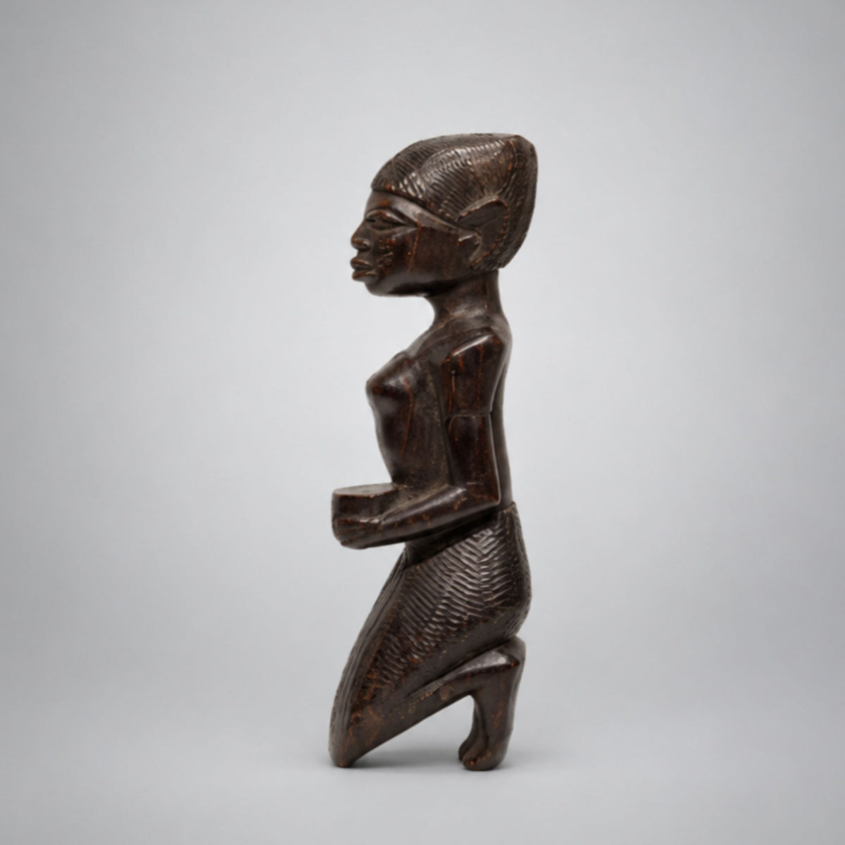 A dark brown carved wooden figurine of a kneeling female figure in profile, shown against a neutral light gray background.