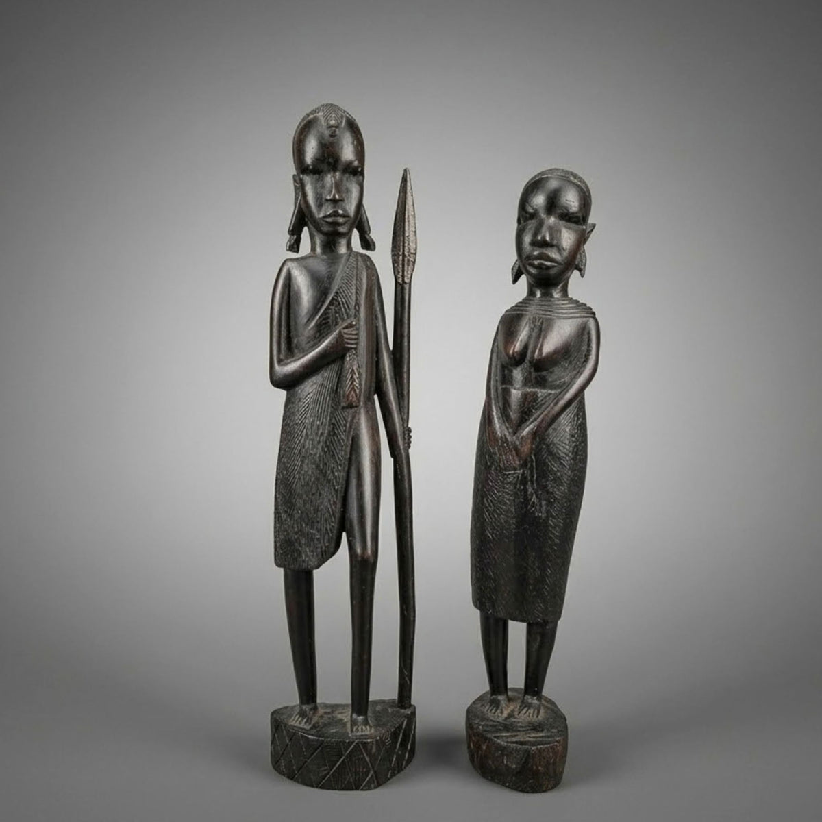 Two dark carved wooden African-style figurines standing on round bases against a smooth gray gradient background. One figure holds a spear, and both have elongated heads and textured clothing details.