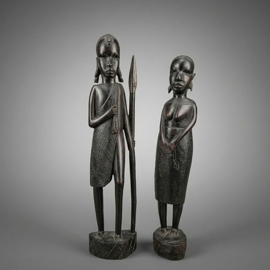 Two dark carved wooden African-style figurines standing on round bases against a smooth gray gradient background. One figure holds a spear, and both have elongated heads and textured clothing details.