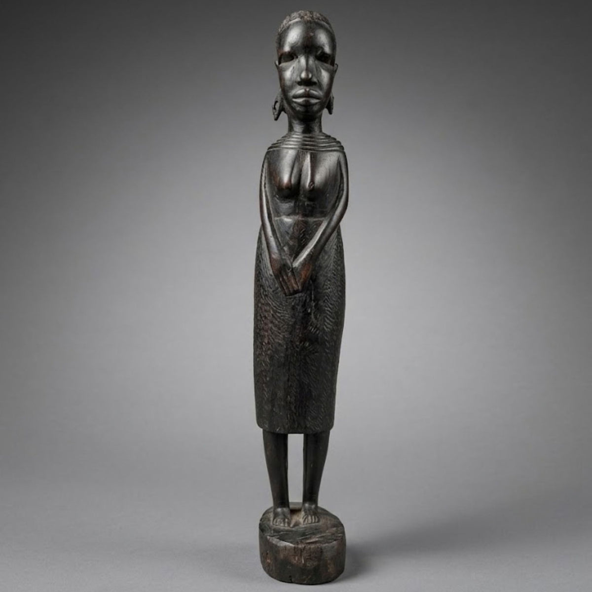 A dark carved wooden statue of a standing female maasai figure with a stylized face, shown against a neutral gray studio background.