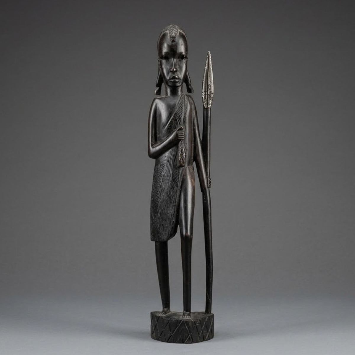 A dark carved wooden statue of a standing figure holding a spear, photographed against a smooth gray studio background.