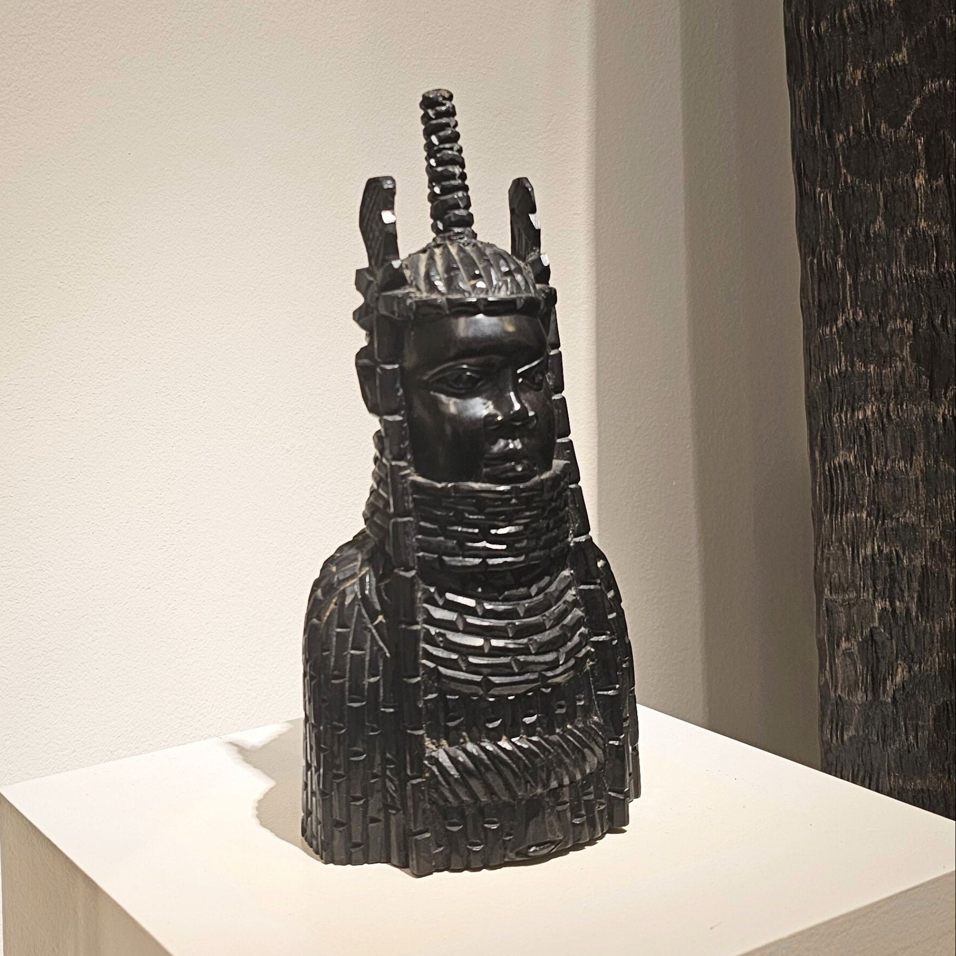 Black sculpture of a figure with intricate details on a white pedestal against a neutral wall.