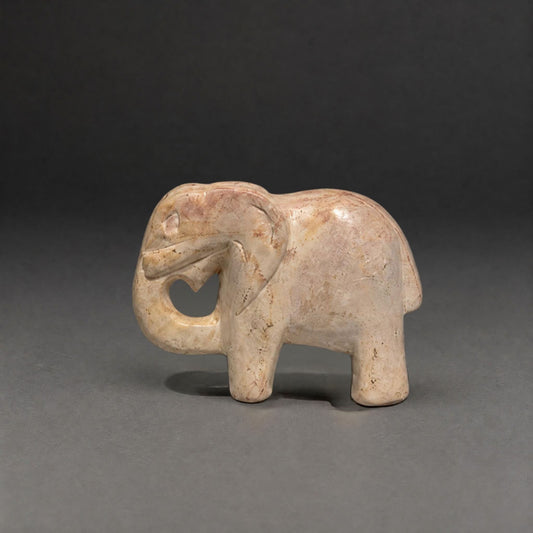 A small carved elephant figurine in a light beige stone or ceramic material with subtle warm brown marbling, standing against a dark gray studio background.