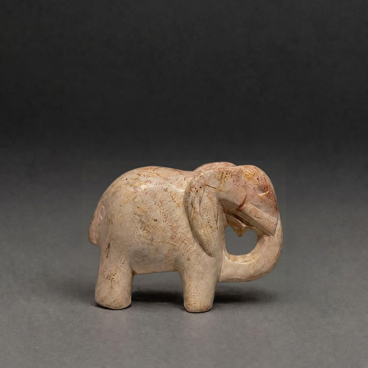 A small carved elephant figurine in a light beige stone or resin with subtle brown marbling, photographed against a dark gray studio background.