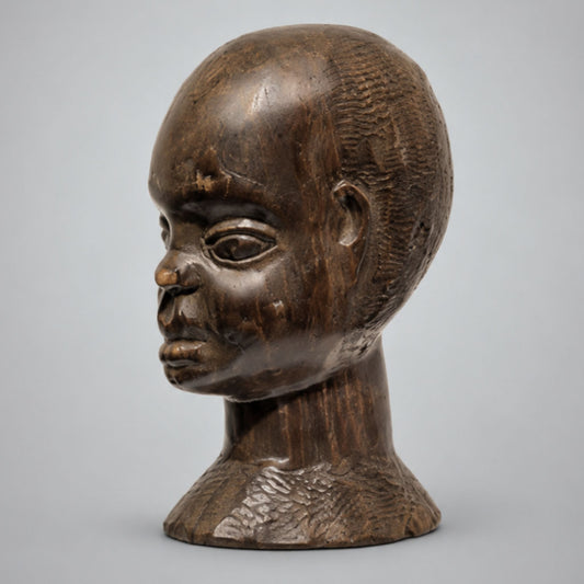 A dark brown carved wooden sculpture of a man's head and neck, featuring detailed textured patterns on the scalp and neck, set against a neutral light gray background.