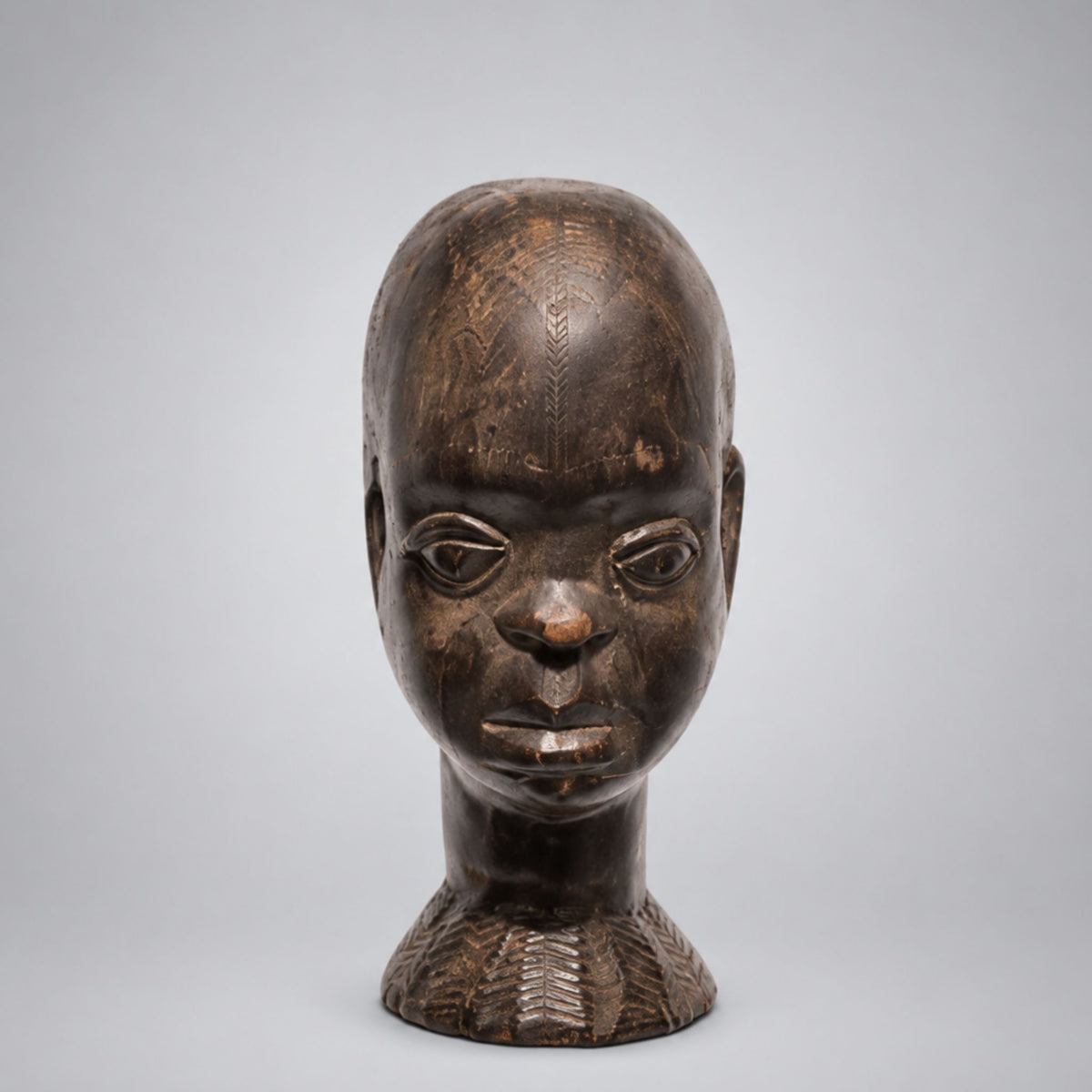 A carved wooden African-style bust sculpture of a man's head and neck, featuring detailed hand-carved patterns and a weathered dark brown finish, set against a neutral light gray background.