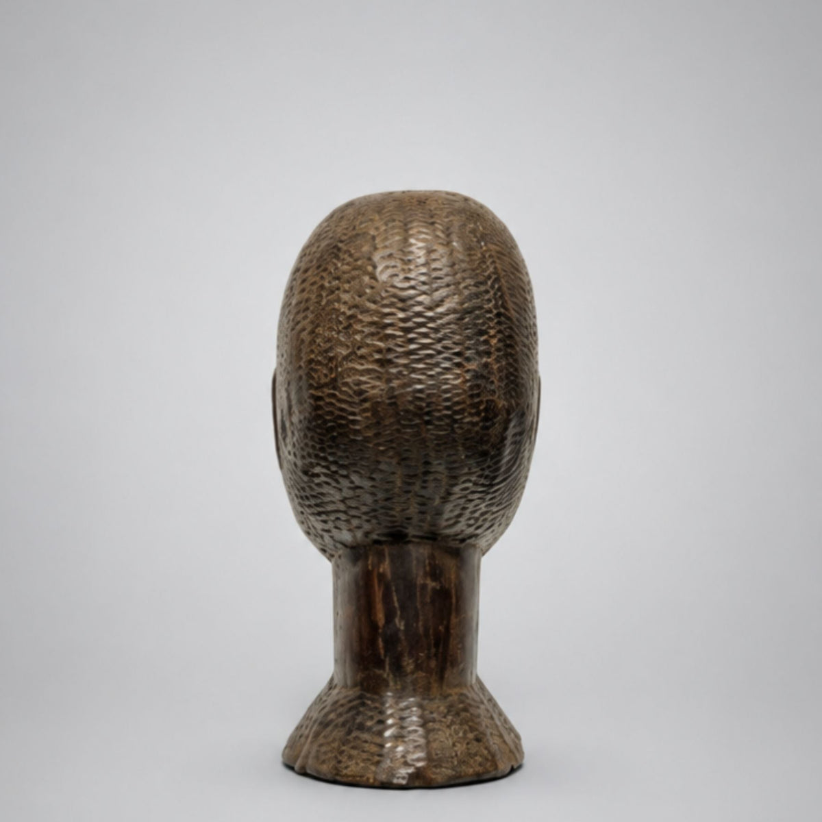 Rear view of A dark brown carved wooden sculpture of a man's head and neck, featuring detailed textured patterns on the scalp and neck, set against a neutral light gray background.