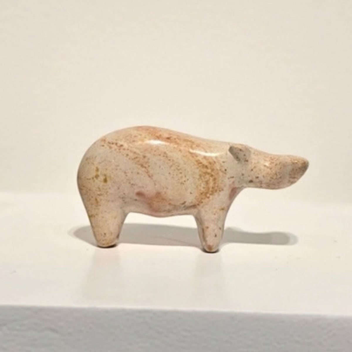 A small carved animal figurine, shaped like a hippo, made from speckled beige stone or ceramic, displayed on a white pedestal against a plain off-white background.