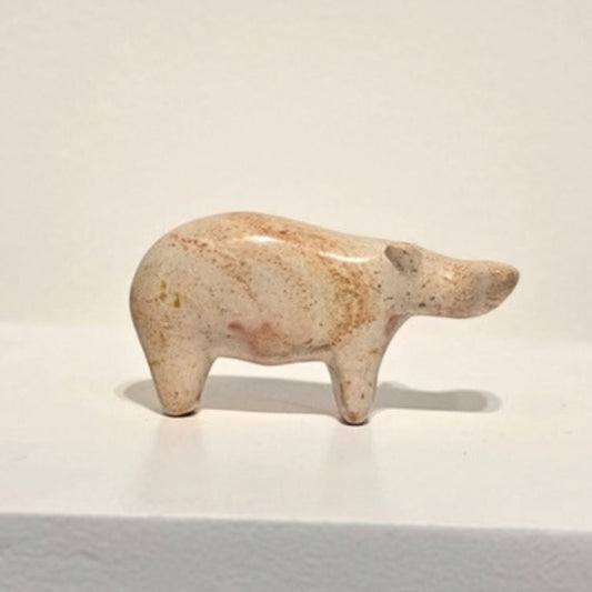 A small carved animal figurine, shaped like a hippo, made from speckled beige stone or ceramic, displayed on a white pedestal against a plain off-white background.