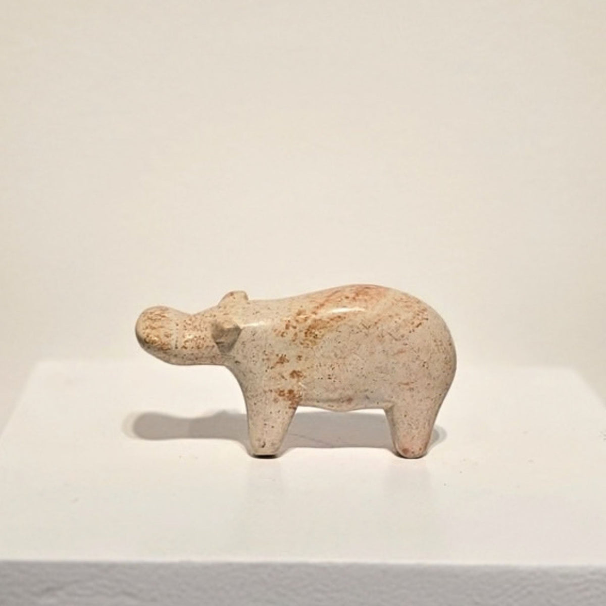 A small hand-carved hippopotamus figurine made of speckled beige stone with brown spots, standing on a white display platform against a plain white background.